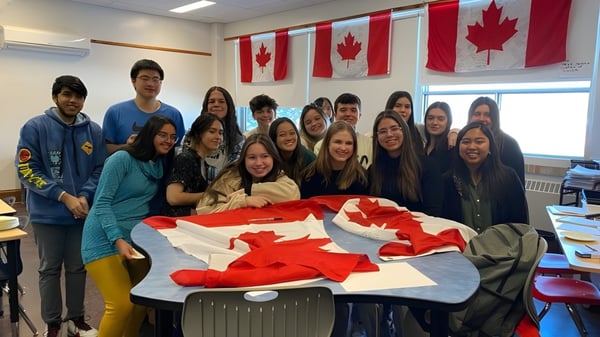 Un grupo de alumnas y alumnos posan juntos en un aula en el campus de St. James Collegiate con banderas canadienses de fondo.