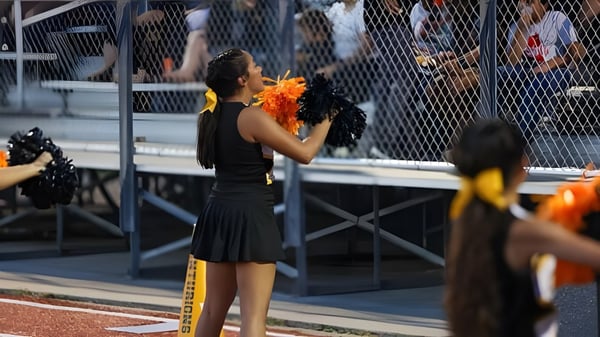 Una persona con un vestido negro sostiene una flor naranja en el escenario de la St. John Paul II High School frente a una audiencia.