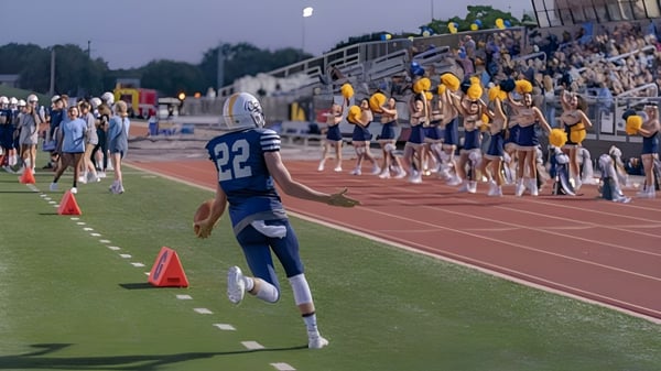 Un jugador de fútbol americano con camiseta azul corre en el campo de la St. John Paul II Catholic High School frente a espectadores en las gradas.