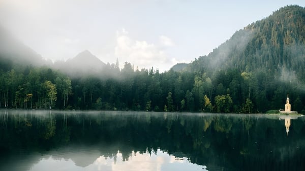 Un lago tranquilo con una pequeña iglesia en la orilla, rodeado de bosques verdes y montañas, cerca de la St. John’s-Kilmarnock School.