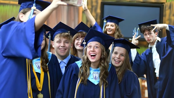 Un grupo de graduadas y graduados de la St. John’s-Ravenscourt School en togas de graduación azules están juntos al aire libre frente a una cerca de madera.