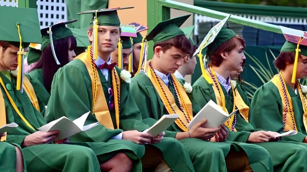 Un grupo de graduadas y graduados de la St. Johnsbury Academy en togas verdes posan con diplomas frente a un fondo colorido.