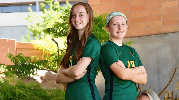 Dos estudiantes de la St. Joseph Catholic High School están en camisetas verdes frente a un edificio de ladrillo con plantas verdes.