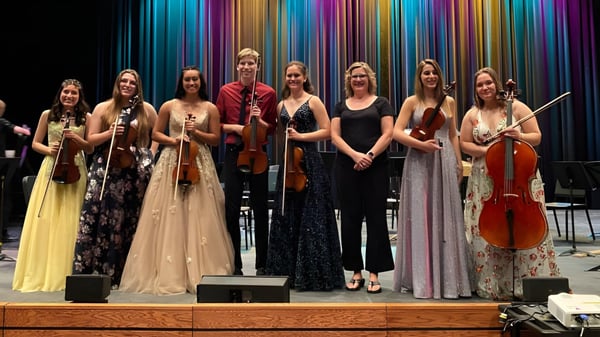 Estudiantes de la St. Joseph High School están vestidos de manera formal con instrumentos musicales en un escenario frente a un fondo colorido.