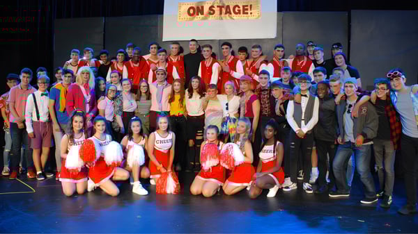 Un grupo de estudiantes de la St. Joseph’s Secondary School está en disfraces coloridos en un escenario con un cartel de On Stage! en el fondo.