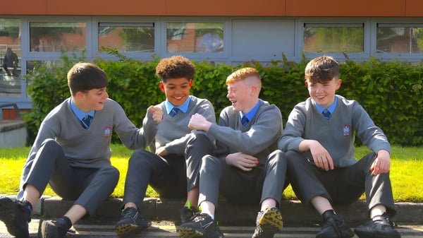 Cuatro estudiantes de la St. Joseph's Secondary School están sentados juntos en un banco frente a un edificio con grandes ventanas.