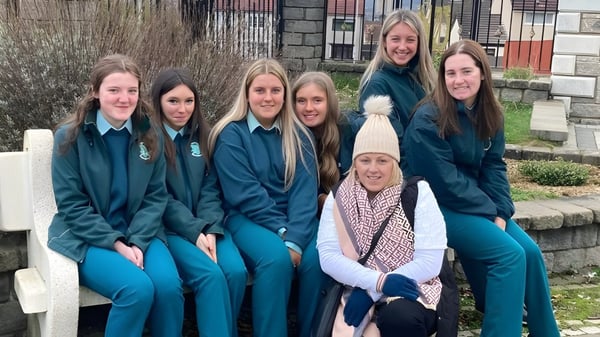 Un grupo de alumnas está frente a una pared de piedra y una cerca de madera en el terreno de la St. Louis Secondary School.