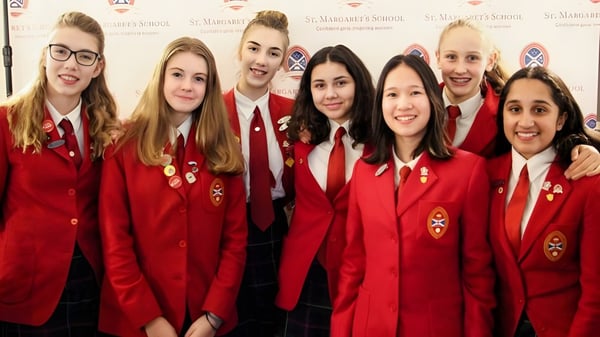 Un grupo de estudiantes de la St. Margaret’s School posan en uniformes rojos frente a un fondo de logo.