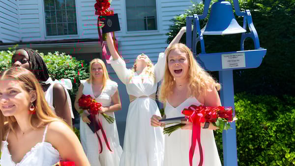 Alumnas de St. Margaret's School en vestidos blancos con flores rojas frente a un edificio de madera blanca.