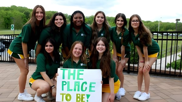Un grupo de estudiantes de la St. Mark High School está con un cartel 'The Place to Be' frente a un parque con árboles y una cerca.