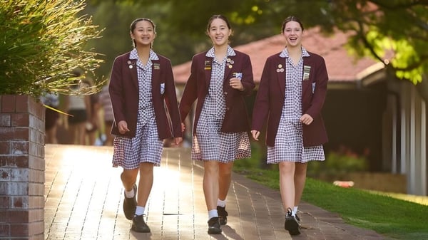 Tres estudiantes de la St. Mary's Anglican Girls' School caminan por un camino entre áreas verdes y edificios de ladrillo.