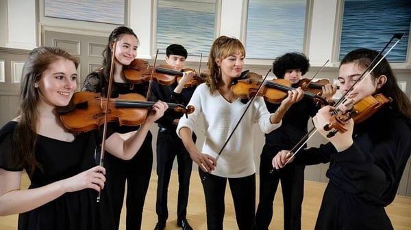 Estudiantes de la St. Mary’s Music School tocan juntos el violonchelo en una sala con grandes ventanas y vista a un cuerpo de agua.