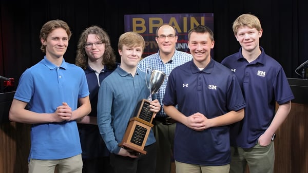 Un grupo de seis estudiantes está con un trofeo en el campus de St. Mary's School frente a un cartel de Brain.