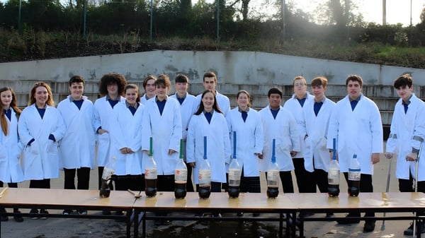 Un grupo de estudiantes en batas de laboratorio blancas están en el terreno de St. Michael's College frente a una pared de concreto con árboles al fondo.