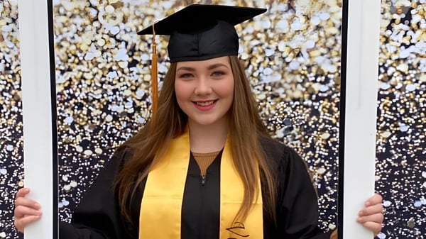 Una joven en toga de graduación está frente a un fondo brillante en el campus de St. Michael's Regional High.
