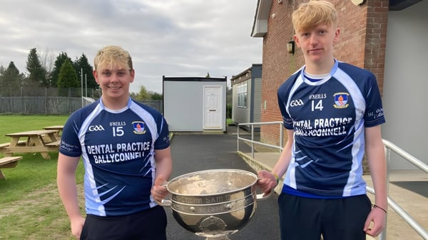 Dos estudiantes del St. Mogues College sostienen un trofeo frente a un edificio de ladrillo y árboles.