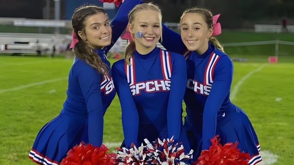 Tres alumnas de la St. Paul Catholic High School posan en uniformes de animadoras con pompones en el campo deportivo de noche.