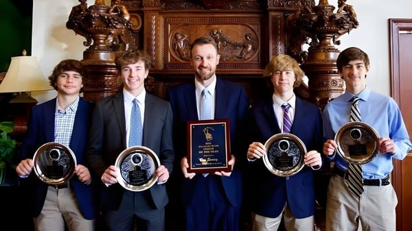 Un grupo de hombres en trajes sostiene premios frente a una pared de madera decorada en el recinto de la St. Paul High School.