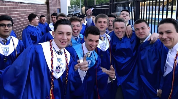 Un grupo de estudiantes en togas de graduación azules posan frente a un edificio de ladrillo en el campus de la St. Peter Catholic High School.