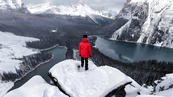 Una persona con chaqueta roja está en un acantilado nevado con vista a un lago congelado y montañas cubiertas de nieve cerca de la St. Pius X High School.
