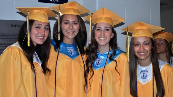 Un grupo de alumnas de la St. Rose High School está en túnicas y birretes amarillos frente a un fondo neutral.