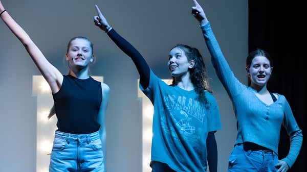 Tres alumnas de St. Teresa’s College muestran expresiones faciales expresivas y levantan los brazos frente a un fondo oscuro.