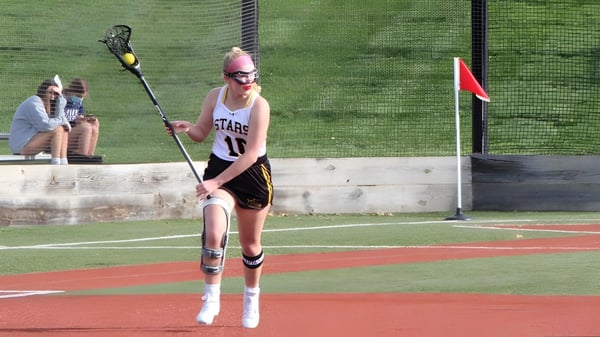 Una persona en camiseta rosa está en el campo de béisbol de St. Teresa’s College frente a hierba y suelo rojo.