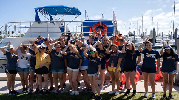 Estudiantes de la St. Thomas Aquinas High School posan en uniformes deportivos uniformes frente a una marina con barcos.