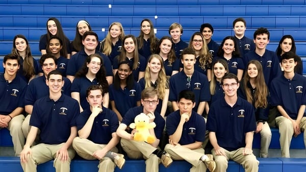 Un grupo de estudiantes de la St. Thomas Aquinas High School posan frente a una pared azul en camisetas oscuras uniformes.
