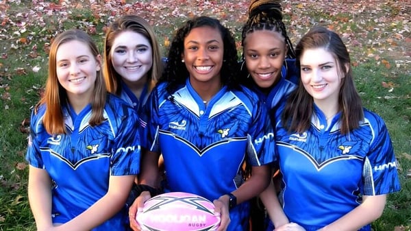 Un grupo de cinco estudiantes de la St. Vincent's Secondary School posan en un uniforme deportivo azul en un campo con coloridas hojas de otoño.