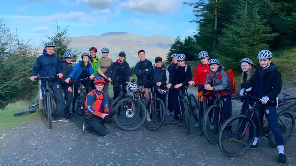 Un grupo de estudiantes de Stamford Endowed Schools está vestido con ropa de ciclista en un camino del bosque con pinos y montañas al fondo.