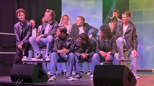 Un grupo de alumnas y alumnos está sentado en el escenario de la Stepaside Educate Together Secondary School frente a un colorido fondo de escenario.