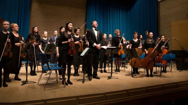 Una gran orquesta de la Stephen Perse Foundation toca junta en el escenario frente a un telón azul.