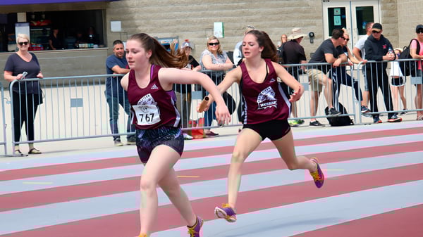 Dos estudiantes de la Stratford District Secondary School corren en la pista durante una competición con espectadores al fondo.