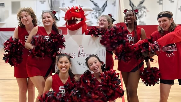 Un grupo de animadoras de la Stuart Hall School posan en uniformes rojos y negros con pompones y un gran disfraz de mascota roja en un gimnasio.
