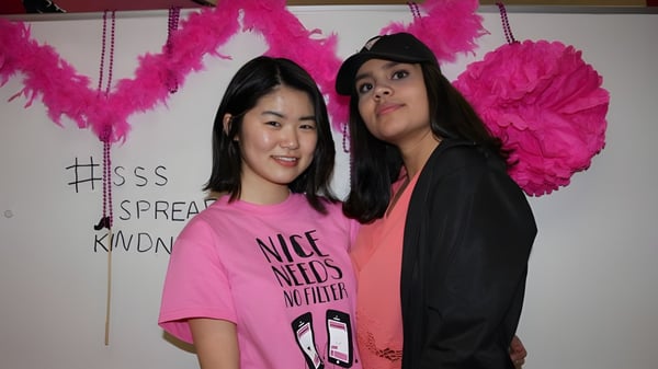 Dos estudiantes de la Summerland Secondary School posan frente a una pared con grandes decoraciones de plumas rosas.