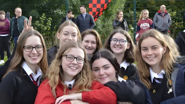 Un grupo de alumnas de la Sutton Park School está juntas en un área parecida a un parque con árboles y una bandera a cuadros rojo y blanco de fondo.