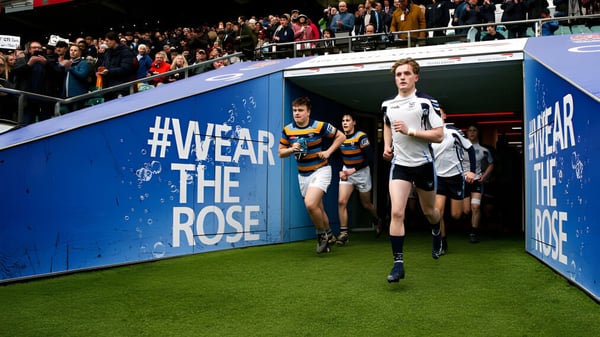 Estudiantes de la Sutton Valence School caminan por un campo frente a un gran banner de #WearTheRose con espectadores al fondo.