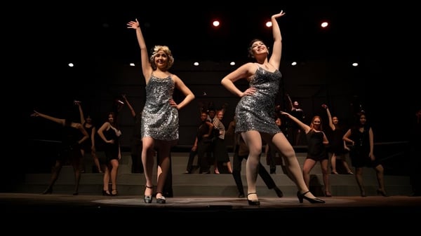 Dos mujeres en vestidos plateados están en el escenario durante una actuación de Tabor Academy con estudiantes bailando al fondo.