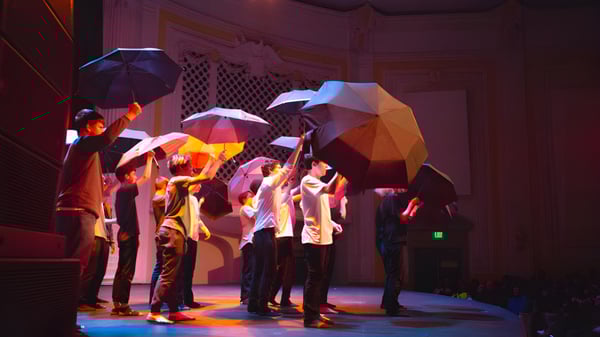 Un grupo de estudiantes de Tacoma Public Schools sostiene paraguas coloridos en un escenario con iluminación dramática.