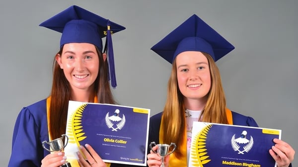 Dos graduadas de la Taumarunui High School llevan ropa de graduación y sostienen sus diplomas frente a un fondo gris.