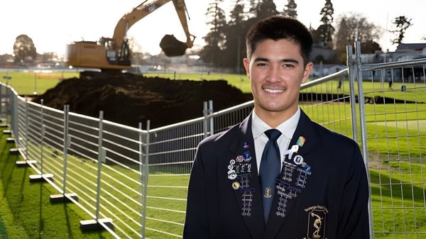 Un estudiante del Tauranga Boy's College sonríe con uniforme frente a un campo deportivo con maquinaria de construcción al fondo.