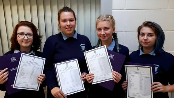 Cuatro estudiantes en uniforme escolar sostienen certificados frente a una pared con persianas verticales en el campus del Templemichael College.