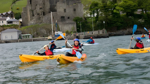Estudiantes del Templemichael College reman en coloridos kayaks en un cuerpo de agua con un castillo histórico al fondo.