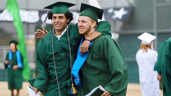 Dos graduados de la Templeton Secondary School se abrazan en togas verdes frente a otros graduados.