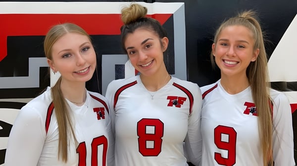 Tres estudiantes de la Terry Fox Secondary School llevan camisetas deportivas blancas con números rojos frente a un fondo rojo y negro.