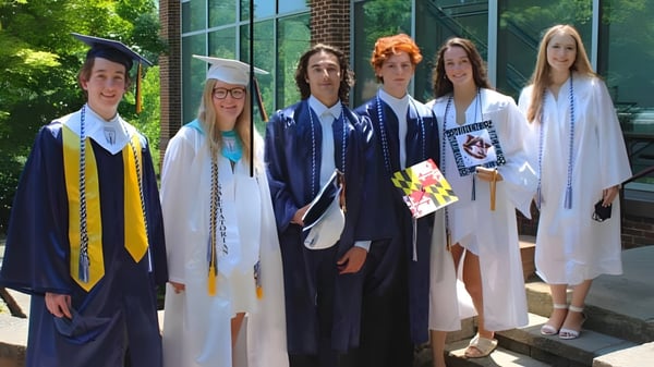 Un grupo de graduados de The Calverton School está en vestimenta académica frente a un edificio de ladrillo con grandes ventanas y plantas.