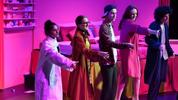 Un grupo de estudiantes de The Friends’ School lleva túnicas coloridas en una sala con iluminación rosa-violeta.