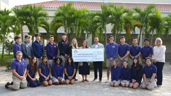 Un grupo de estudiantes de The King's Academy con uniformes azules sostiene un gran cheque frente a un edificio con palmeras.