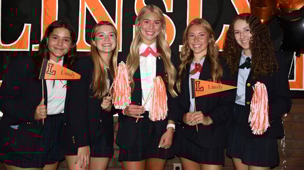 Un grupo de estudiantes de The Linsly School está en uniformes negros con acentos rosas frente a un gran cartel de Lindy y sostiene pompones rosas.
